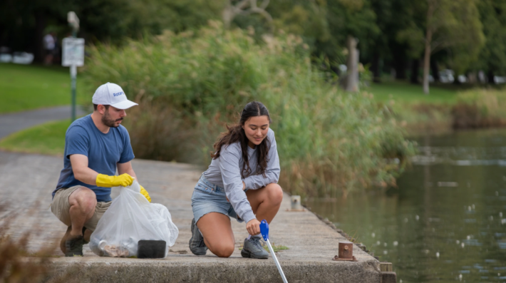 Two individuals on a pier extending to a water body, picking out plastic rubbish from the water body 
