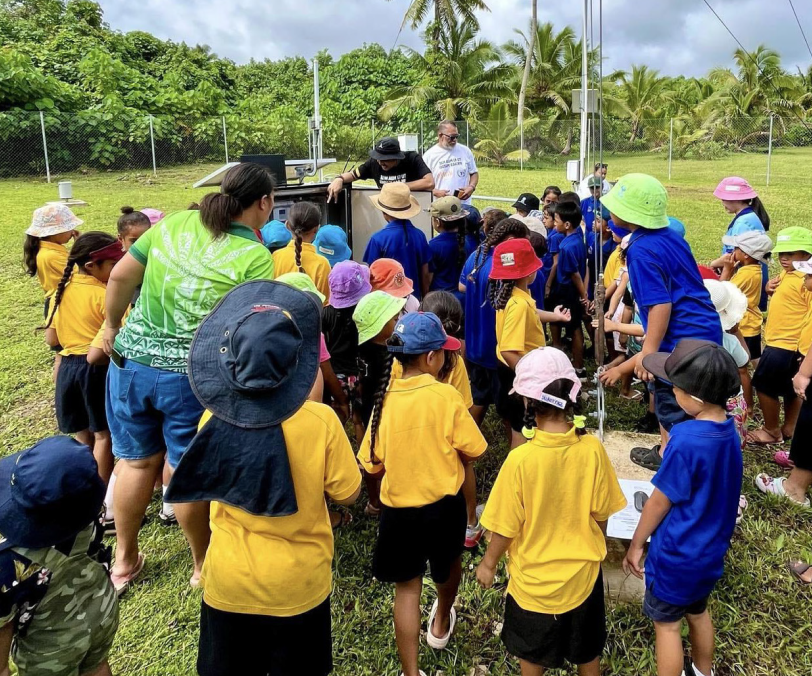 Children gather around some equipment in a field with palm trees in the background 