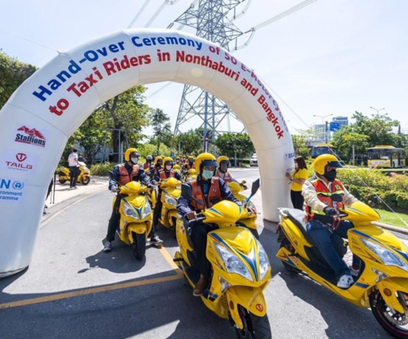 Several yellow electric motorcycles driving down the street and passing under a banner