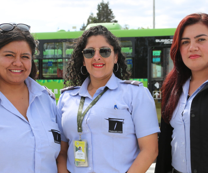 Three women in uniform posing for a photo and smiling with a green bus behind them  